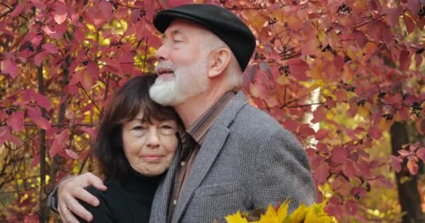 Blisko starsza para, brodaty mąż z siwymi włosami przytulający smutną żonę z bukietem jesiennych liści, pocieszający radosny żart uśmiechający się i śmiejący. Romantyczne relacje spacerujących seniorów. - Materiał filmowy, wideo