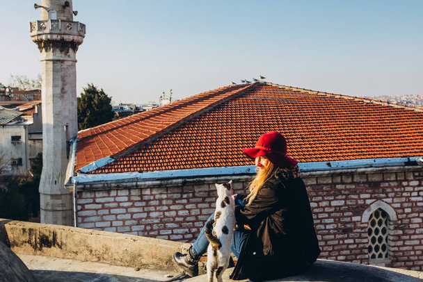 Blond woman stroking a cat on the roof with a view to the mosque, Istanbul, Turkey. Girl in a hat sits on the roof in Istanbul, sunny autumn day. Traveler girl walks through winter Istanbul. - Photo, Image