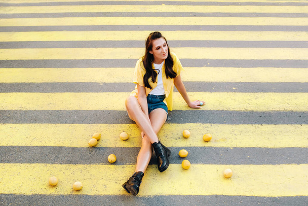 an unusual girl in a yellow shirt, shorts and black shoes with lemons is sitting on a pedestrian crossing in the city. The lemon mood - 写真・画像