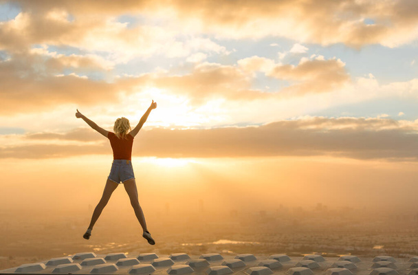 Success and uplifting. Happy woman feeling good and fulfilled jumping in the air celebrating life on the rooftop of a skyscraper overlooking the city view during a beautiful sunrise. - Photo, Image