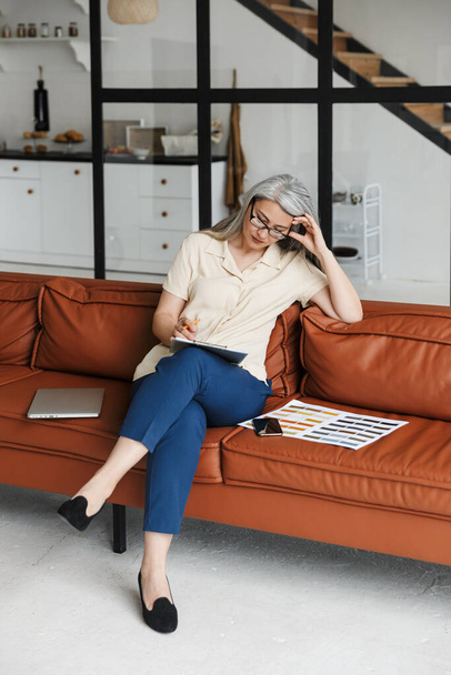 Photo of a serious mature business woman designer sitting on sofa near colors catalogue indoors at home while writing notes. - Foto, imagen