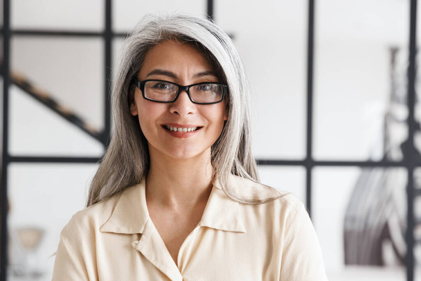Photo of happy gray-haired asian woman wearing eyeglasses smiling and looking at camera in living room - Zdjęcie, obraz