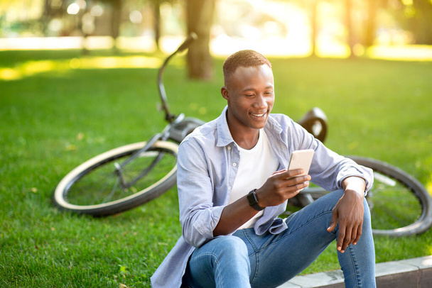 Funky African American man with bicycle browsing internet on his smartphone at urban park - Fotó, kép