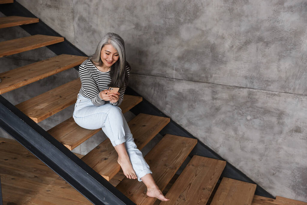 Immagine di donna matura ottimista seduta su gradini al chiuso a casa durante l'utilizzo del telefono cellulare. - Foto, immagini