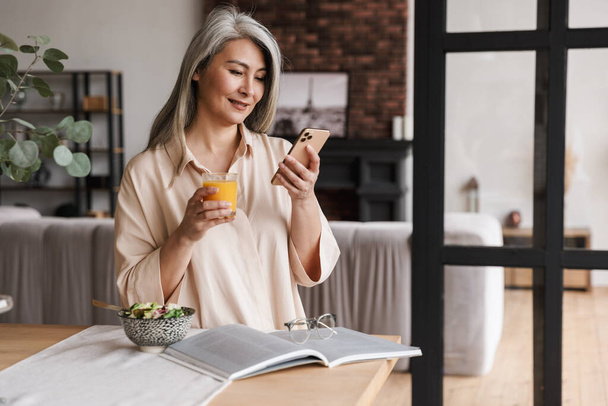 Photo of mature cheerful woman sitting at kitchen indoors at home while chatting by phone and drinking juice. - Photo, Image
