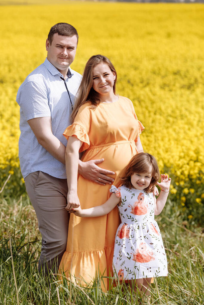 Happy pregnant family with little daughter spending time together in a yellow rapeseed field on summer day. - Photo, Image