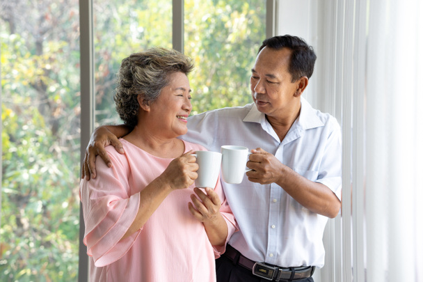 Happy senior couple looking to each other eyes smiling while enjoy drinking their morning coffee at home. Retirement lifestyle concept - Photo, Image