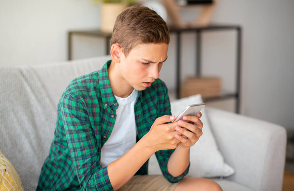Wütender Teenager blickt auf Smartphone-Bildschirm, Wohnzimmereinrichtung - Foto, Bild