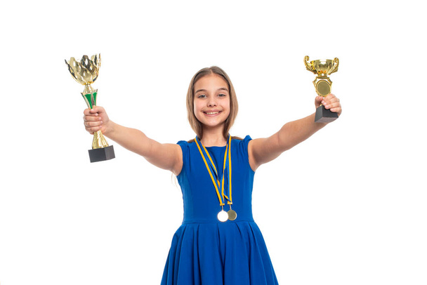 Ritratto di mezza lunghezza di una giovane adolescente felice e di successo che indossa un vestito blu tiene in mano una tazza dorata, premi - medaglie appese al collo. Il concetto di un'infanzia felice. - Foto, immagini