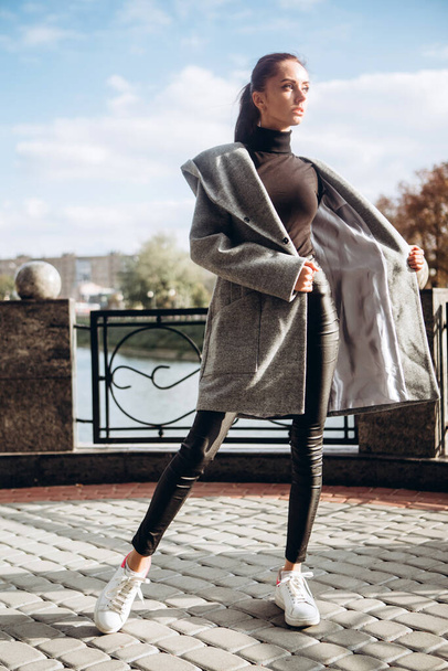 girl in a warm coat outdoors. model in outerwear. a brunette in a long gray coat walks along the embankment of the river. beautiful girl in a fashionable casual coat. spring autumn collection. - Valokuva, kuva