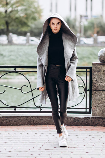 girl in a warm coat outdoors. model in outerwear. a brunette in a long gray coat walks along the embankment of the river. beautiful girl in a fashionable casual coat. spring autumn collection. - Photo, image