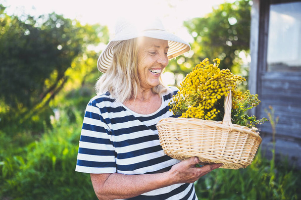 Szczęśliwy uśmiechnięty starsza pani w słomkowym kapeluszu o zabawy pozowanie w letnim ogrodzie z kwiatami w koszyku. Rolnictwo, ogrodnictwo, rolnictwo, emeryci. Hodowla roślin organicznych w gospodarstwie.  - Zdjęcie, obraz
