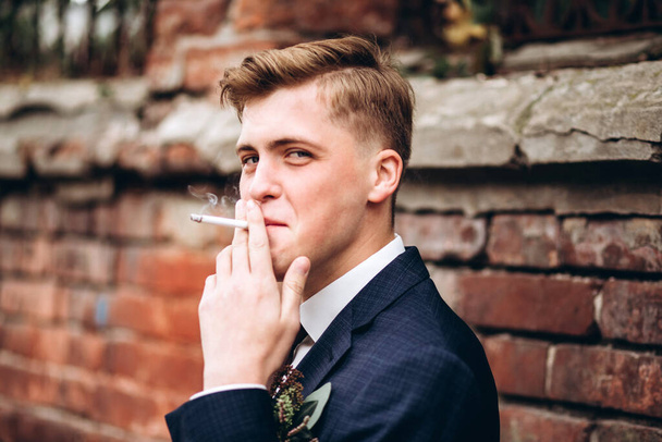 スーツを着た残酷な新郎とネクタイはタバコを吸う。金髪の男が旧市街を歩いている。喫煙や悪い習慣。レンガの壁の背景に若い男性実業家。笑顔の男のポート. - 写真・画像