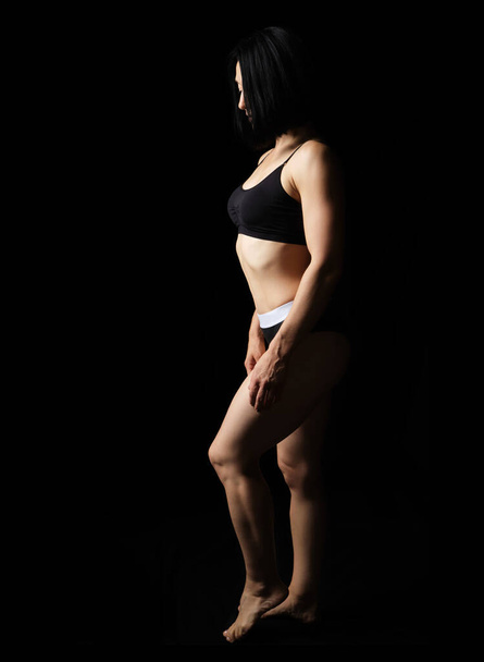 adulto caucasico aspetto donna in un reggiseno nero e bikini si trova su uno sfondo nero, bel corpo muscolare, taglio di capelli corto - Foto, immagini