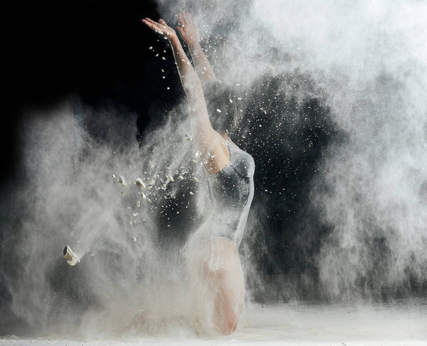 bella donna caucasica in un body nero con una figura sportiva sta ballando in una nuvola bianca di farina su uno sfondo nero - Foto, immagini