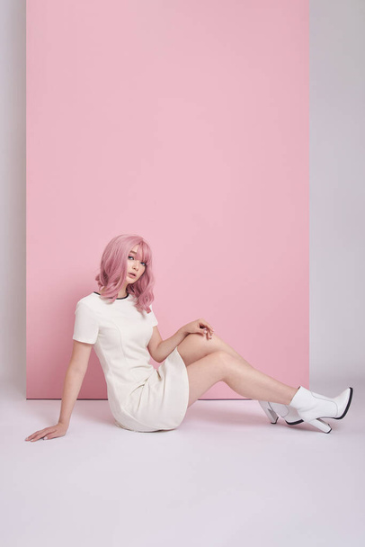 Kaunis tyttö pitkät vaaleanpunaiset hiukset istuu lattialla, hiusten värjäys. Söpö nainen vaaleanpunaisella taustalla lyhyessä valkoisessa mekossa. Värilliset hiukset, täydellinen kampaus - Valokuva, kuva