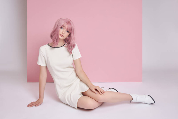 Beautiful girl with long pink hair sitting on the floor, hair coloring. Cute woman on a pink background in a short white dress. Colored hair, perfect hairstyle - Foto, afbeelding