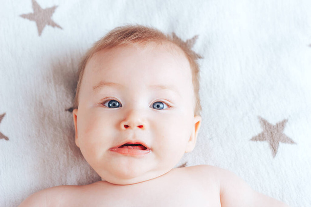 Fénykép egy rémült csecsemőről, egy újszülöttről kék szemekkel és szőke hajjal, amint egy világos takarón fekszik barna csillagokkal.. - Fotó, kép