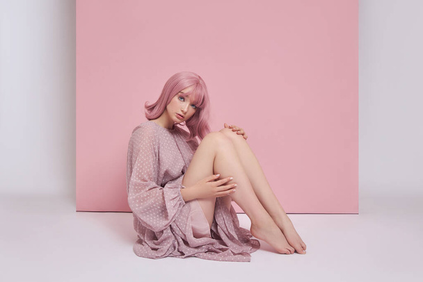 Woman with dyed pink hair in a long dress is sitting on the floor. Portrait of a girl with hair coloring at the pink wall. Perfect hairstyle and hair styling - Photo, Image