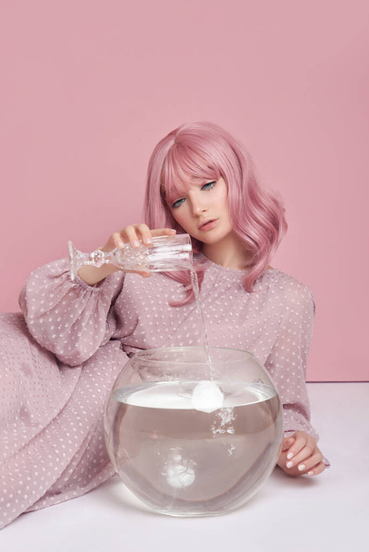 Girl with dyed pink hair in long dress sitting on the floor with a round aquarium. Portrait woman with hair coloring at pink wall. Perfect hairstyle and hair styling - 写真・画像