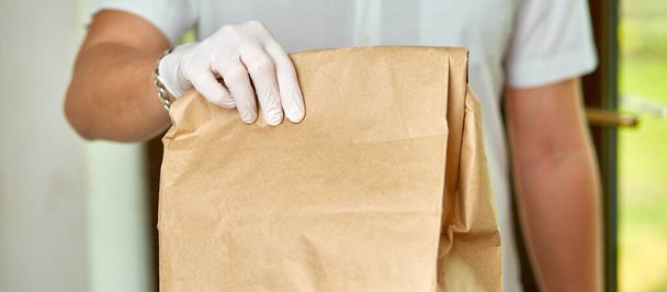 Kurier, kurier w medycznych rękawiczkach lateksowych bezpiecznie dostarcza zakupy online w brązowych papierowych torbach do drzwi podczas epidemii koronawirusa, COVID-19. Zostań w domu, bezpieczna koncepcja.. - Zdjęcie, obraz