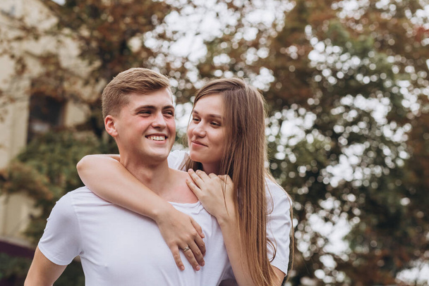 Happy young couple walks the streets of the city and hold hands. guy and girl in white t-shirts and jeans outdoors. Teenagers cuddling against the backdrop of an autumn tree. Couple close-up portrait - 写真・画像