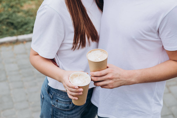 Giovane coppia felice cammina per le strade della città e beve caffè da una tazza di cartone. ragazzo e ragazza in t-shirt bianche e jeans all'aperto. Adolescenti coccole sullo sfondo del paesaggio urbano. - Foto, immagini