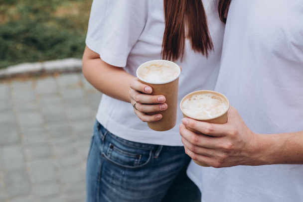 Ευτυχισμένο νεαρό ζευγάρι περπατά στους δρόμους της πόλης και πίνει καφέ από ένα χάρτινο κύπελλο. άντρας και κορίτσι με λευκά μπλουζάκια και τζιν έξω. Οι έφηβοι αγκαλιάζονται με φόντο το αστικό τοπίο. - Φωτογραφία, εικόνα