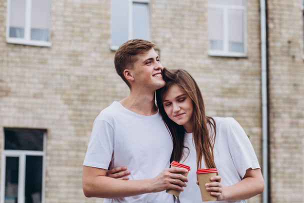 Happy young couple walks the streets of the city and drinks coffee from a cardboard cup. guy and girl in white t-shirts and jeans outdoors. Teenagers cuddle against the backdrop of the cityscape. - Photo, Image