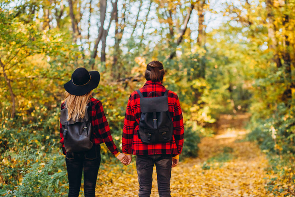 young couple walks through the autumn forest holding hands. Stylishly dressed guy and girl. The guy and the girl in red jackets on background of yellow forest. Happy young couple in love at the park - Photo, Image