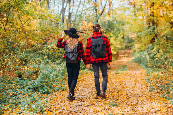 Genç çift sonbahar ormanında el ele tutuşarak yürüyor. Güzel giyimli bir erkek ve kız. Adam ve kırmızı ceketli kız sarı ormanın arka planında. Parkta aşık mutlu genç bir çift - Fotoğraf, Görsel