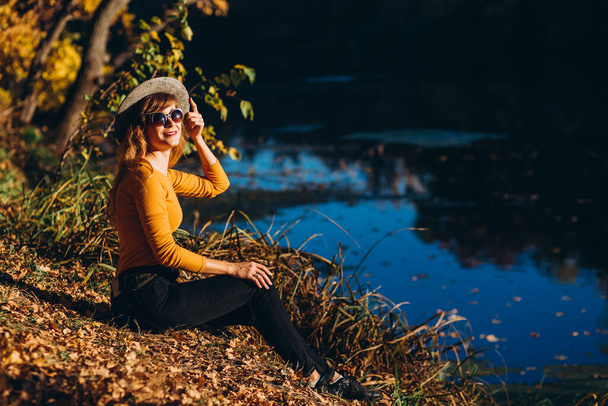 Blondynka w szarym kapeluszu przechodzi przez jesienny las. Wesoła dziewczyna w żółtej bluzie siedzi nad jeziorem w słoneczny jesienny dzień. Kobieta spacerująca po lesie.. - Zdjęcie, obraz