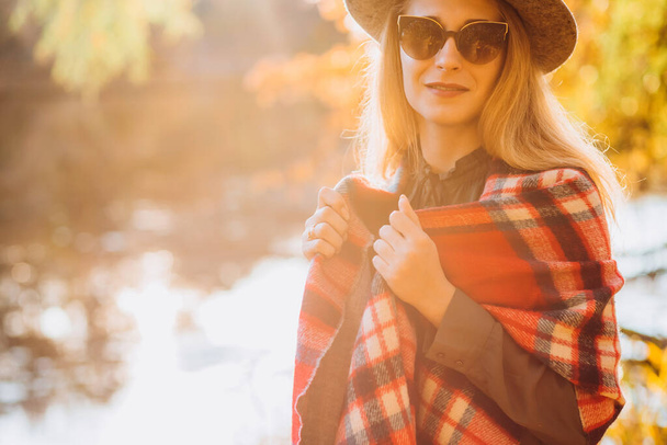 灰色の帽子のブロンドは秋の森を歩く。赤いチェックのスカーフの明るい女の子は黄色の森の背景に立っています。秋には少女のクローズアップ肖像画。ぼやけた背景 - 写真・画像