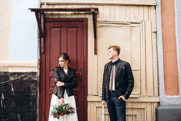 Young happy couple is walking on a city street after rain. Bride and groom with leather jackets outdoors. Autumn wedding. - Фото, зображення