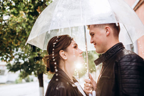 Young happy couple kiss under a transparent umbrella on a city street. Close-up portrait of man and woman. Loving couple smiling under an umbrella. Sunny autumn day in the city. - Φωτογραφία, εικόνα