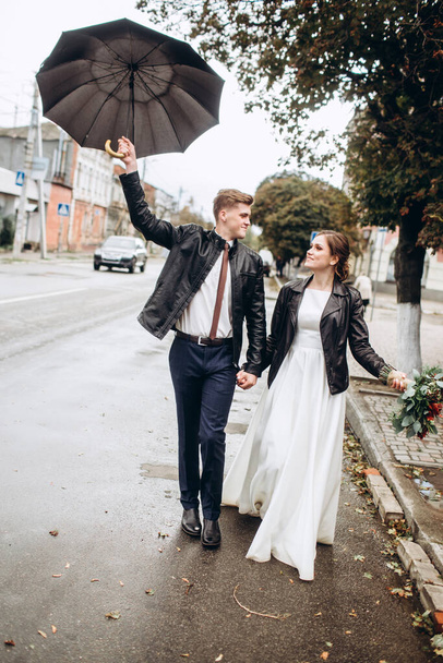 Young happy couple is walking on a city street after rain. Close-up portrait of a man and woman with an umbrella. Loving couple smiling outdoors. A man walks down the street and waving an umbrella - Photo, Image