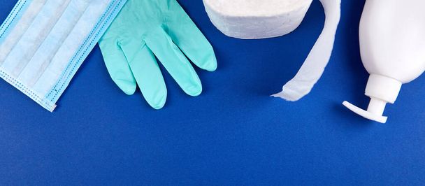 Coronavirus pandemie, crisis winkelen concept. Flat lay van wc-papier gestapelde broodjes, medicijnmasker, handschoenen, hand ontsmettingsmiddel flessen op een blauwe achtergrond. Kopieerruimte. - Foto, afbeelding