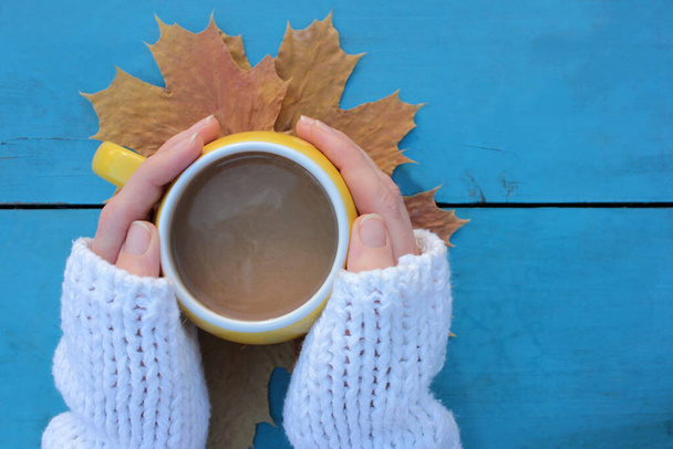 Female hands in white sweater holding yellow cup of coffee on autumn leaves and blue wooden table background. Cozy autumn mood. Top view, copy space, selective focus - Photo, Image