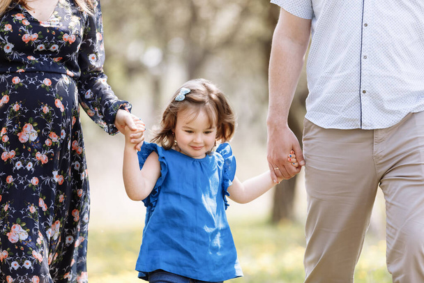 Widok na malucha. Mamo, ojciec trzymają się za ręce córka cieszyć się naturą i chodzić w parku wiosennym. Młoda rodzina spędza razem czas na wakacjach, na świeżym powietrzu. Pojęcie urlopu rodzinnego - Zdjęcie, obraz