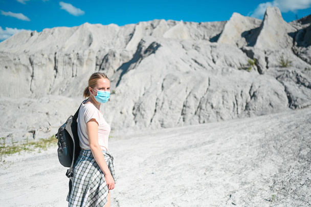 若いブロンドの女性は白い山に向かい、 Covid 19の予防のために顔保護マスクを着用して歩く。リュック付きの若い学生旅行者の観光客。コロナウイルスと国内観光の概念. - 写真・画像