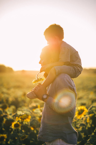 Vater mit Kind in einem Feld blühender Sonnenblumen bei Sonnenuntergang. Vatertag. Seitenansicht - Foto, Bild