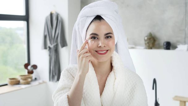 Conceito de cuidados com a pele. Retrato de jovem feliz em roupão de banho branco com toalha na cabeça aplicando creme no rosto, olhando para a câmera, passando a manhã no banheiro, fazendo procedimento de cuidados com a pele - Foto, Imagem
