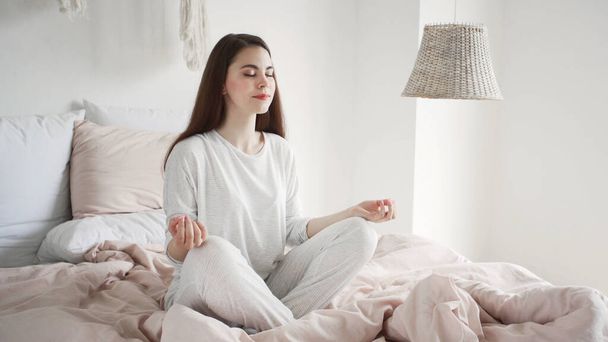 Koncepcja zdrowia psychicznego. Spokojne i marzycielskie młoda kobieta w piżamie siedzi w pozycji medytacji na łóżku w przytulnej sypialni, spędzając rano w domu, odpoczywając i uśmiechając miło - Zdjęcie, obraz