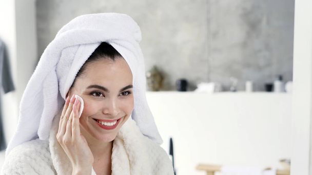 Portret van een vrolijke jonge volwassen vrouw in witte badjas en handdoek op het hoofd brengen ochtend in de badkamer, met behulp van wattenschijfje, het verwijderen van make-up, glimlachen, staande tegen kopieerruimte achtergrond - Foto, afbeelding