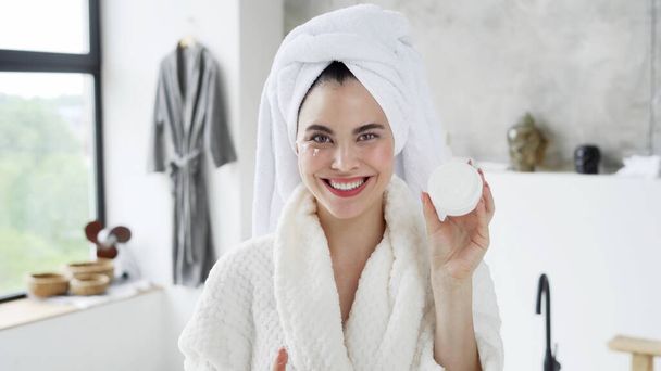 Concept of skincare, anti aging and beauty procedure. Portrait of happy young female in white bathrobe with towel on head showing container with face cream at camera, spending morning at bathroom - Foto, immagini
