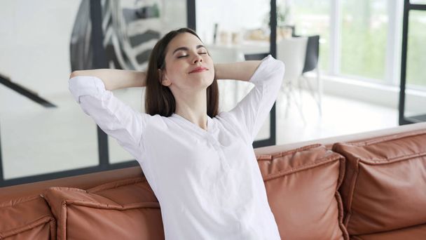 Snící a klidná mladá žena sedící na gauči v neformálním oblečení, držící se za ruce nad hlavou, zavřené oči a odpočívající v obývacím pokoji. Koncept dovolené a víkendu doma - Fotografie, Obrázek