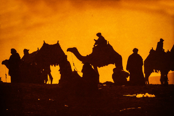 Bir grup deve ve ve binicileri kum tepesine vardıklarında dramatik bulutlu bir çöl günbatımına karşı siluet oluştururlar. Arka planda ufukta uzak bir dağ zirvesi yükseliyor.. - Fotoğraf, Görsel