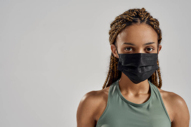 Siyah tıbbi koruyucu maske takmış, gri arka planda tek başına kameraya bakan sportif melez bir kadının portresi. - Fotoğraf, Görsel