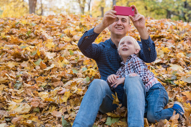 Otec a syn sedí na podzimním listí a fotí na telefonu - Fotografie, Obrázek