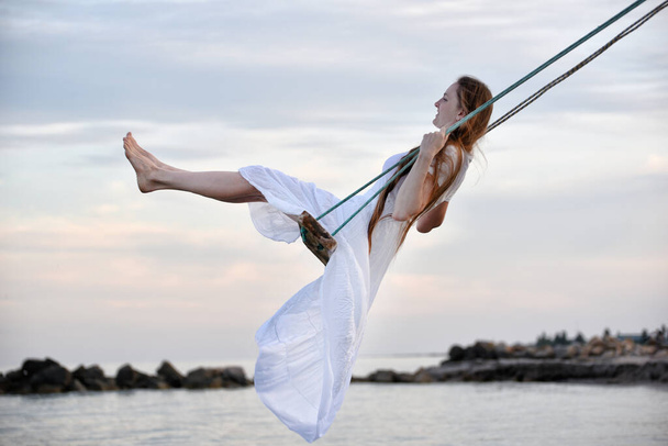Piękna kobieta w białym stroju jeździ na huśtawce liny nad wodą. Wieczór panieński na morzu - Zdjęcie, obraz